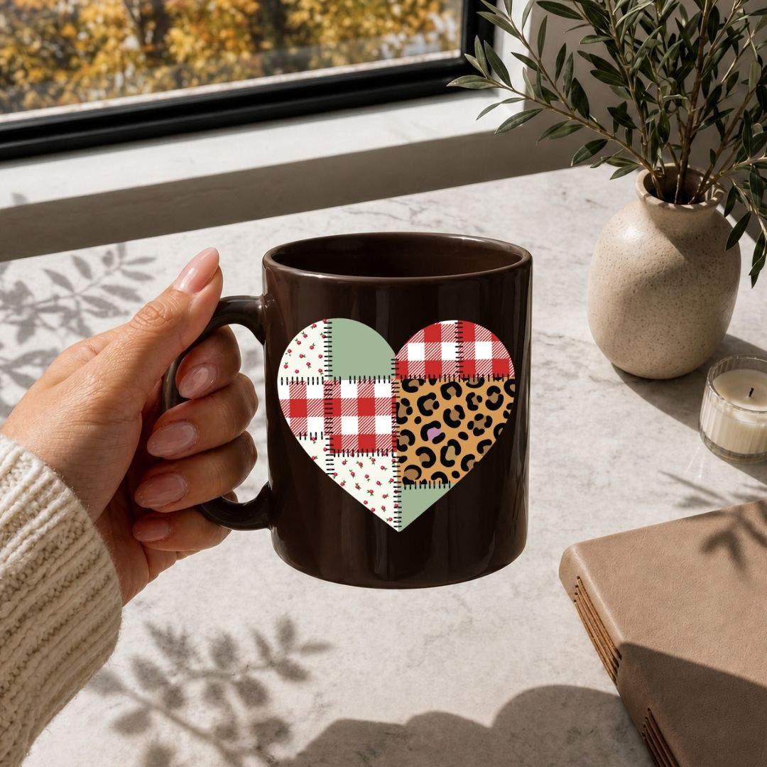 Hand holding a mug with a heart design featuring plaid and leopard patterns on a table with a notebook and candle.