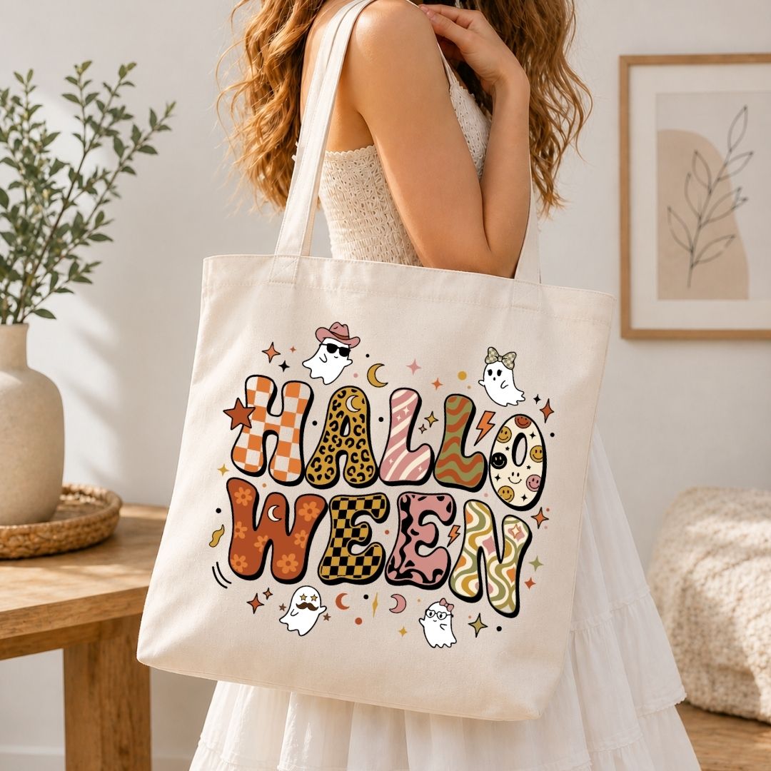 A woman wearing a white dress carries a cream canvas tote bag with a colorful 'HALLOWEEN' graphic featuring ghosts, stars, and patterned letters in an indoor setting.