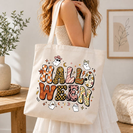 A woman wearing a white dress carries a cream canvas tote bag with a colorful 'HALLOWEEN' graphic featuring ghosts, stars, and patterned letters in an indoor setting.