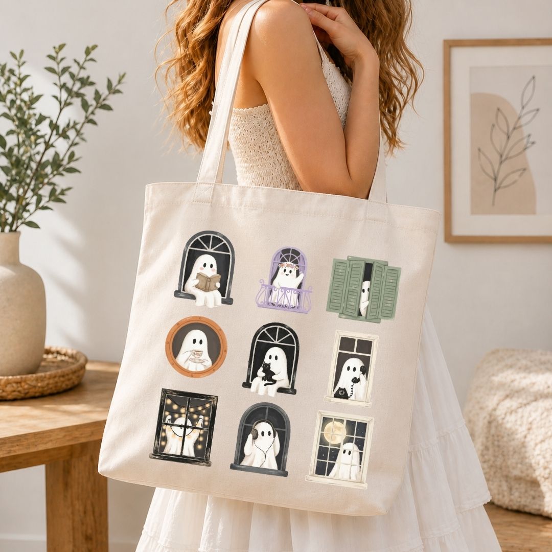 A woman in a white dress carries a cream canvas tote bag decorated with nine cute ghost illustrations in various window poses. The scene is set in a cozy, softly-lit room with a wooden table, potted plant, and framed wall art in the background.