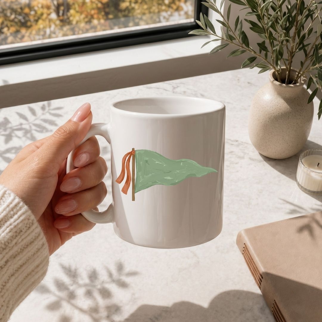 White mug with 'MAMA' text held by a hand on a light surface with a window and plant in the background.