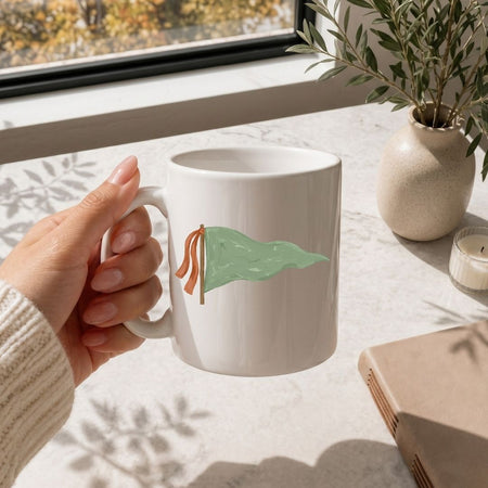 White mug with 'MAMA' text held by a hand on a light surface with a window and plant in the background.