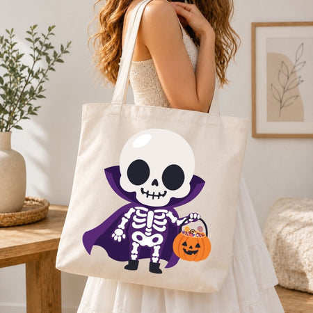 A woman wearing a white sleeveless dress carries a cream canvas tote bag featuring a cartoon skeleton in a purple cape holding an orange jack-o'-lantern bucket. The scene is set in a bright, modern room with a wooden table, potted plant, and framed wall art.