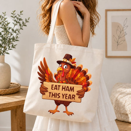 A woman wearing a white dress carries a cream canvas tote bag featuring a cartoon turkey holding a sign that reads 'EAT HAM THIS YEAR' in a cozy indoor setting.