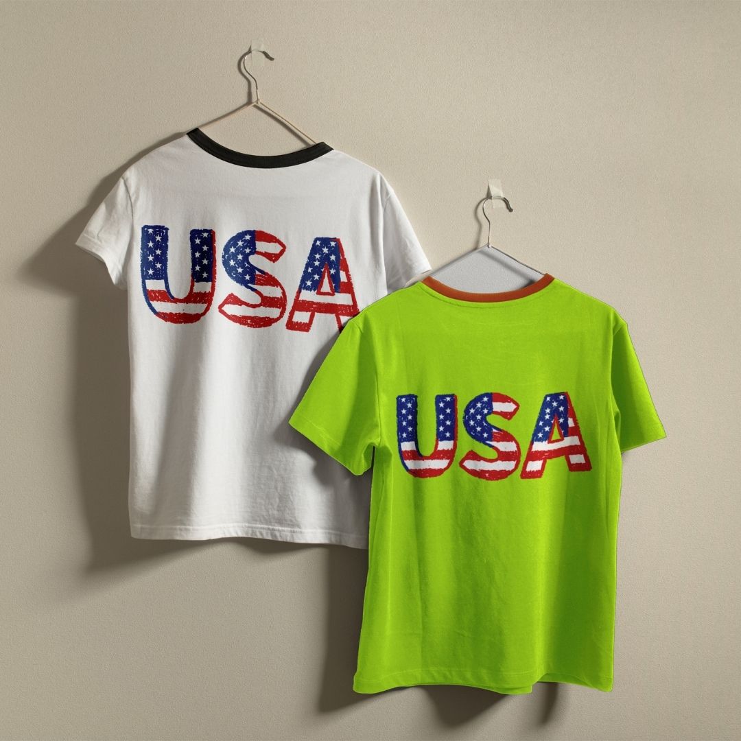Two t-shirts with 'USA' text on hangers against a beige wall.