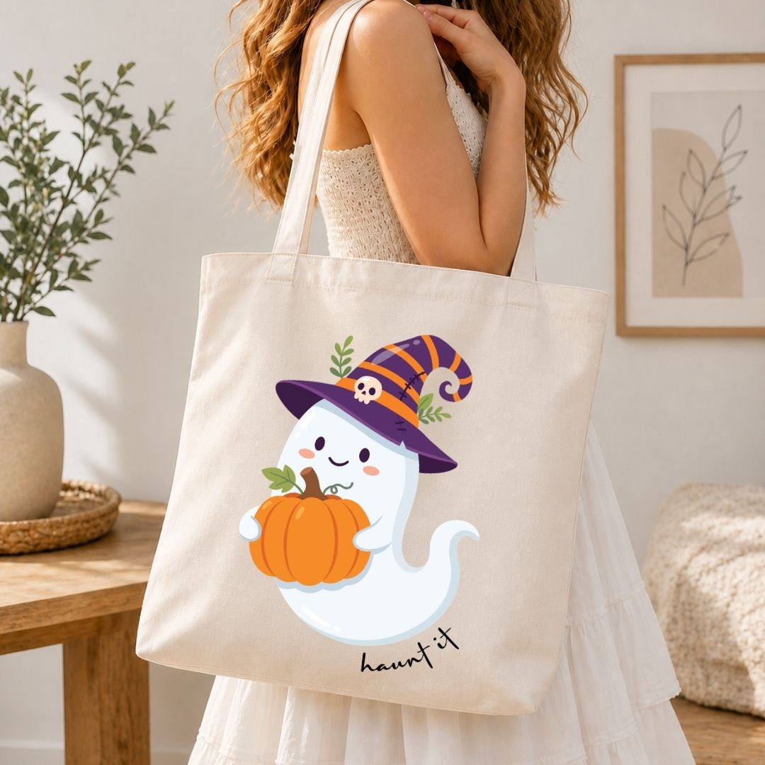 A woman wearing a white dress carries a cream canvas tote bag with a cute cartoon ghost wearing a purple witch hat and holding an orange pumpkin. The tote bag has the word 'haunted' printed in black script at the bottom.
