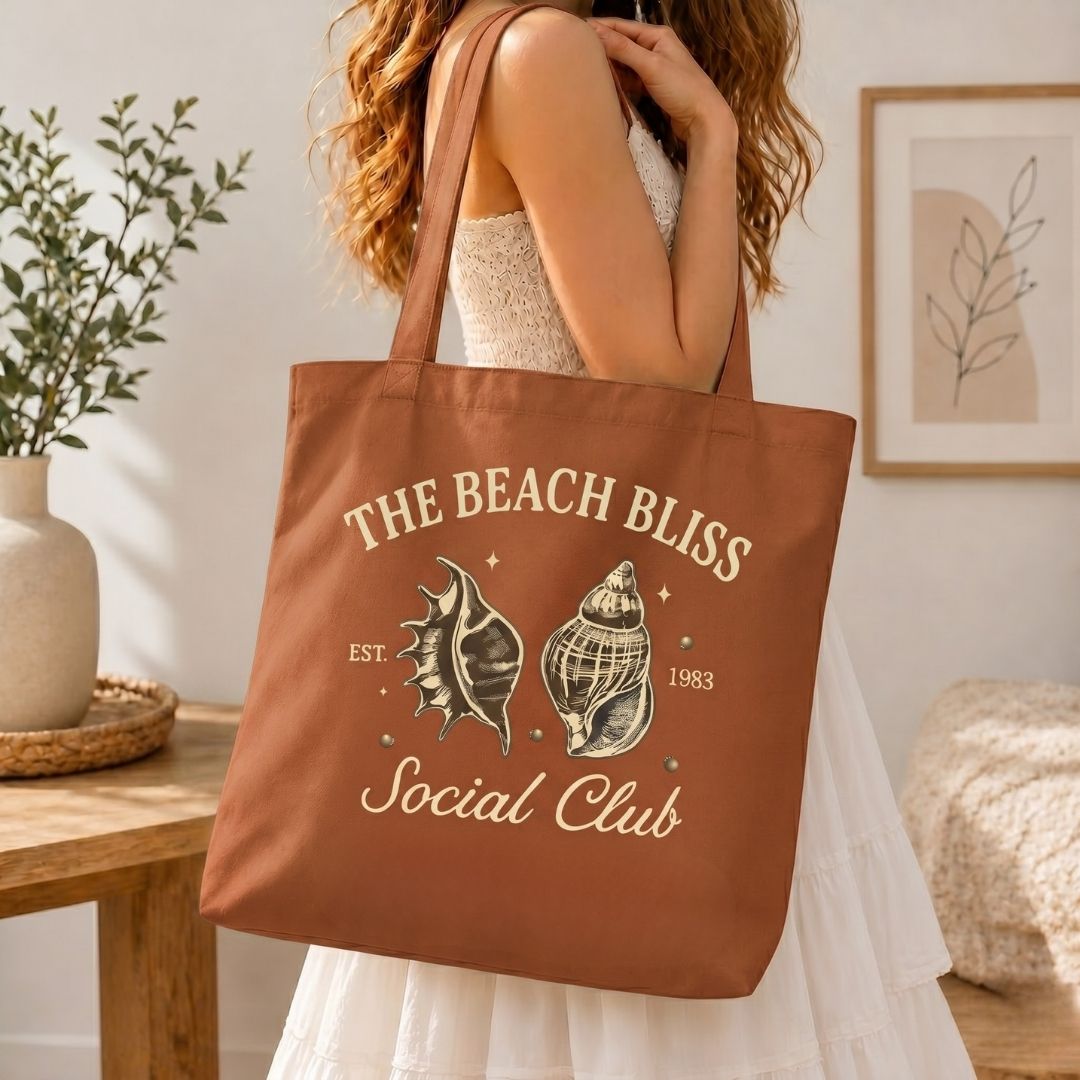 A woman carrying a terracotta rust-colored canvas tote bag over her shoulder, printed with a large vintage seashell graphic and text reading The Beach Bliss Social Club.