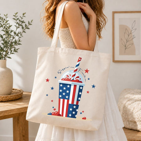 A woman carrying a cream-colored canvas tote bag over her shoulder printed with a large, vibrant 4th of July slushie cup design.