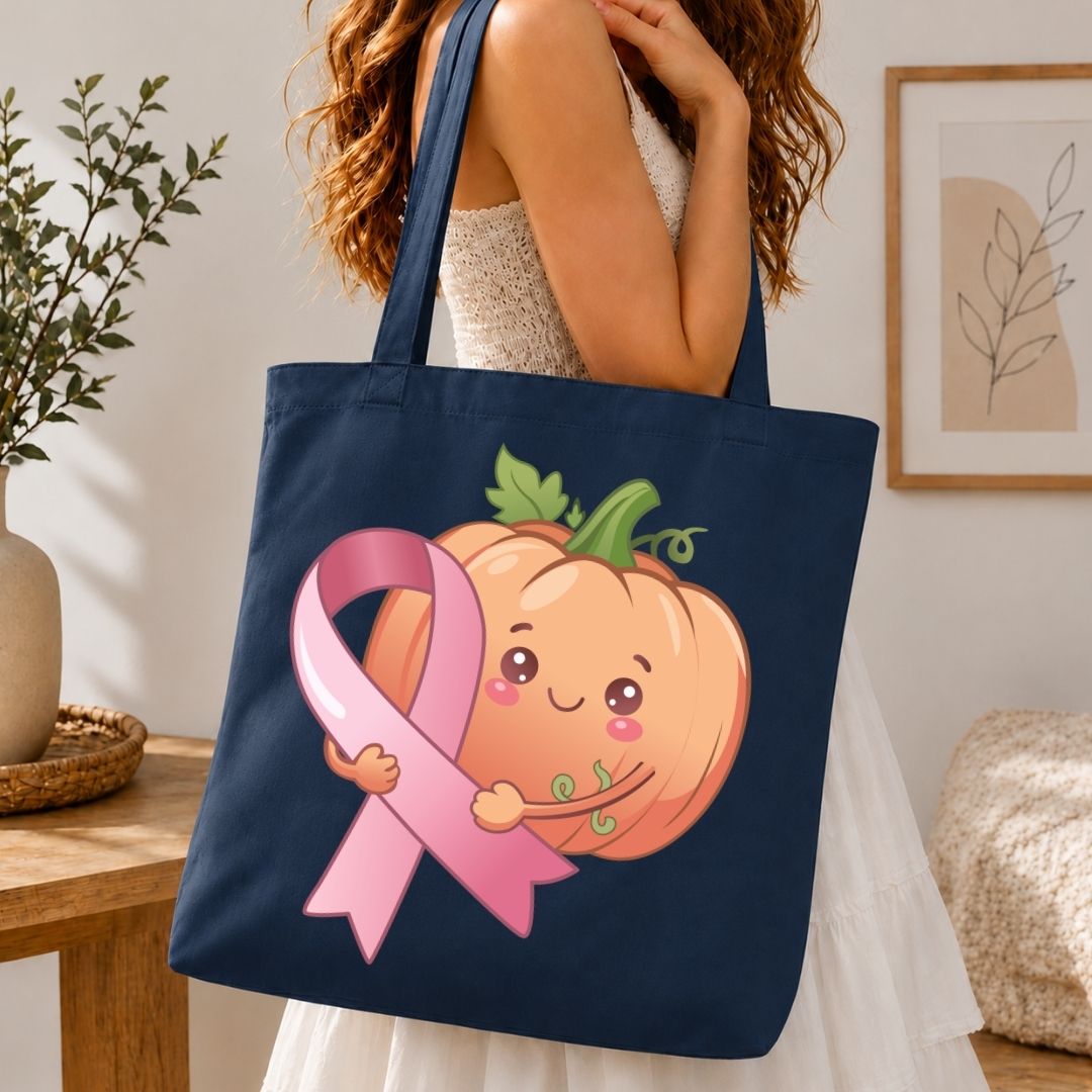 A navy blue canvas tote bag featuring a cute cartoon pumpkin hugging a pink awareness ribbon, carried by a woman in a white dress in a cozy indoor setting.