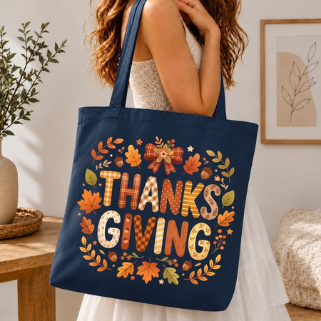 A navy blue canvas tote bag with a 'THANKSGIVING' autumn-themed graphic, carried by a woman in a white dress in a cozy indoor setting.