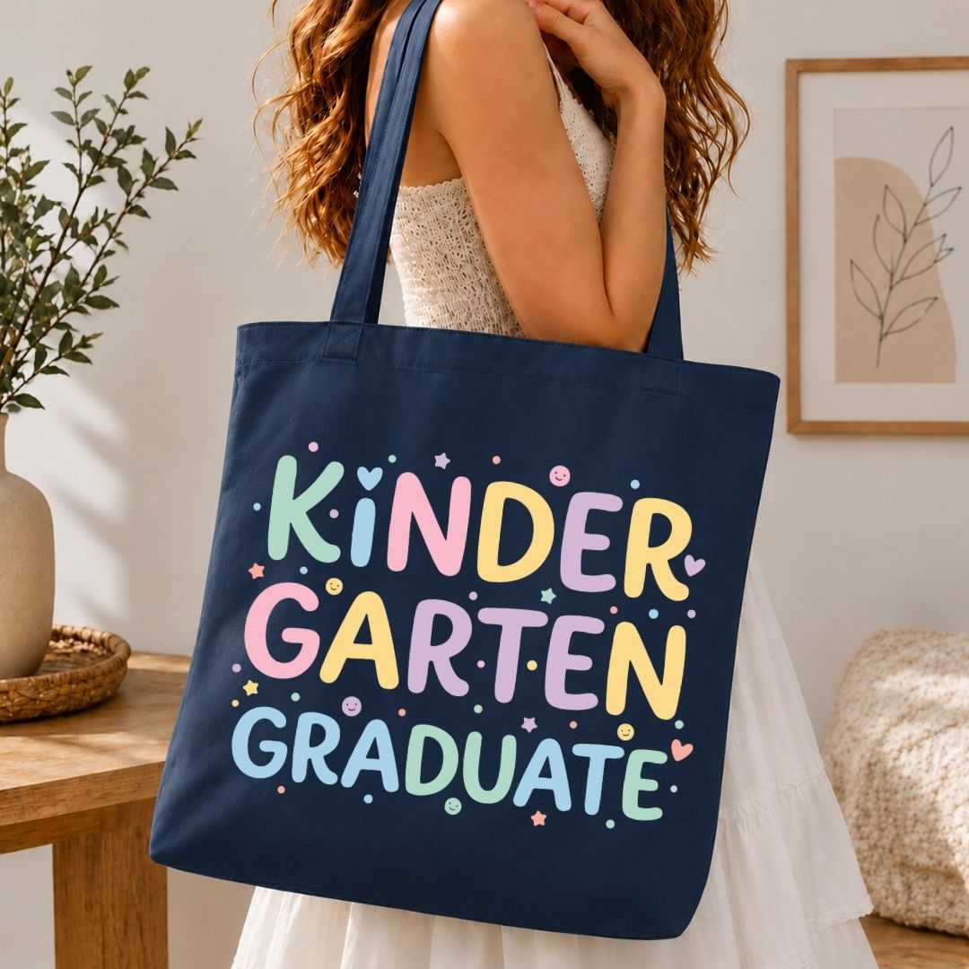 A navy blue canvas tote bag with the words 'KINDER GARTEN GRADUATE' printed in colorful pastel letters, carried by a woman in a white dress in a warm, indoor room setting.