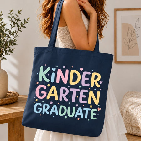 A navy blue canvas tote bag with the words 'KINDER GARTEN GRADUATE' printed in colorful pastel letters, carried by a woman in a white dress in a warm, indoor room setting.