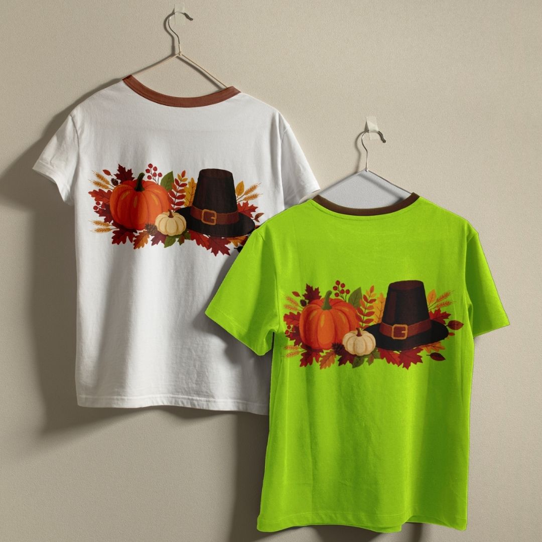 White and green t-shirts with autumn-themed design featuring pumpkins and a pilgrim hat on hangers against a beige background.