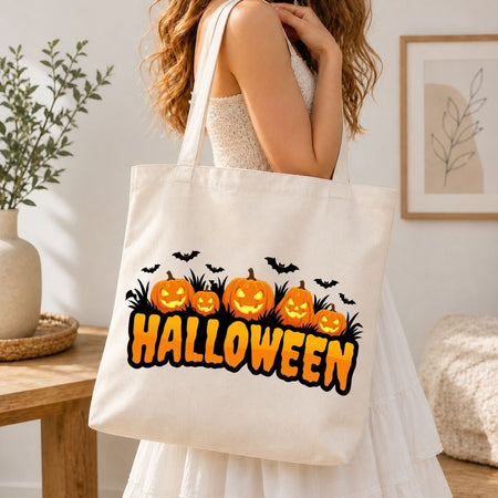 Woman holding a canvas tote bag printed with a Halloween jack-o'-lantern, grass silhouette, and flying bats sublimation graphic.