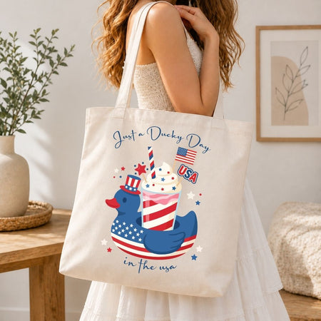 A woman carrying a cream-colored canvas tote bag over her shoulder printed with a large, vibrant 4th of July slushie cup design.
