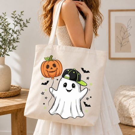 A woman wearing a white dress carries a cream canvas tote bag decorated with a cute cartoon ghost wearing a green and black cap, next to an orange jack-o'-lantern holding a balloon.