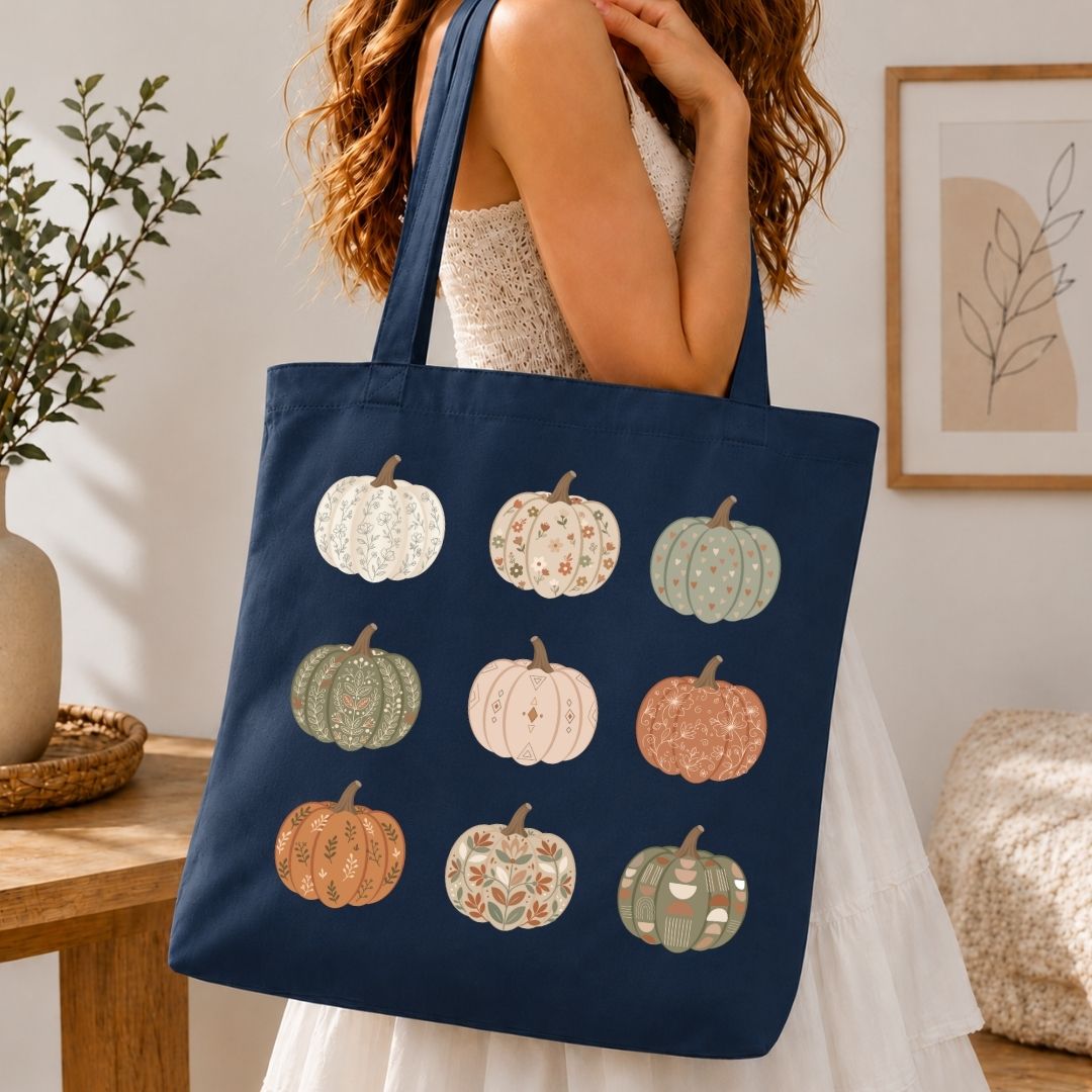 A navy blue canvas tote bag with a pattern of nine autumn pumpkins in various colors, carried by a woman in a white dress in a cozy indoor setting.
