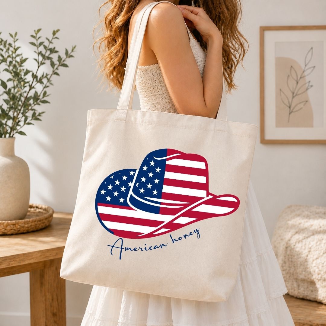 A woman carrying a cream-colored canvas tote bag over her shoulder printed with a large, vibrant American flag cowboy hat design.