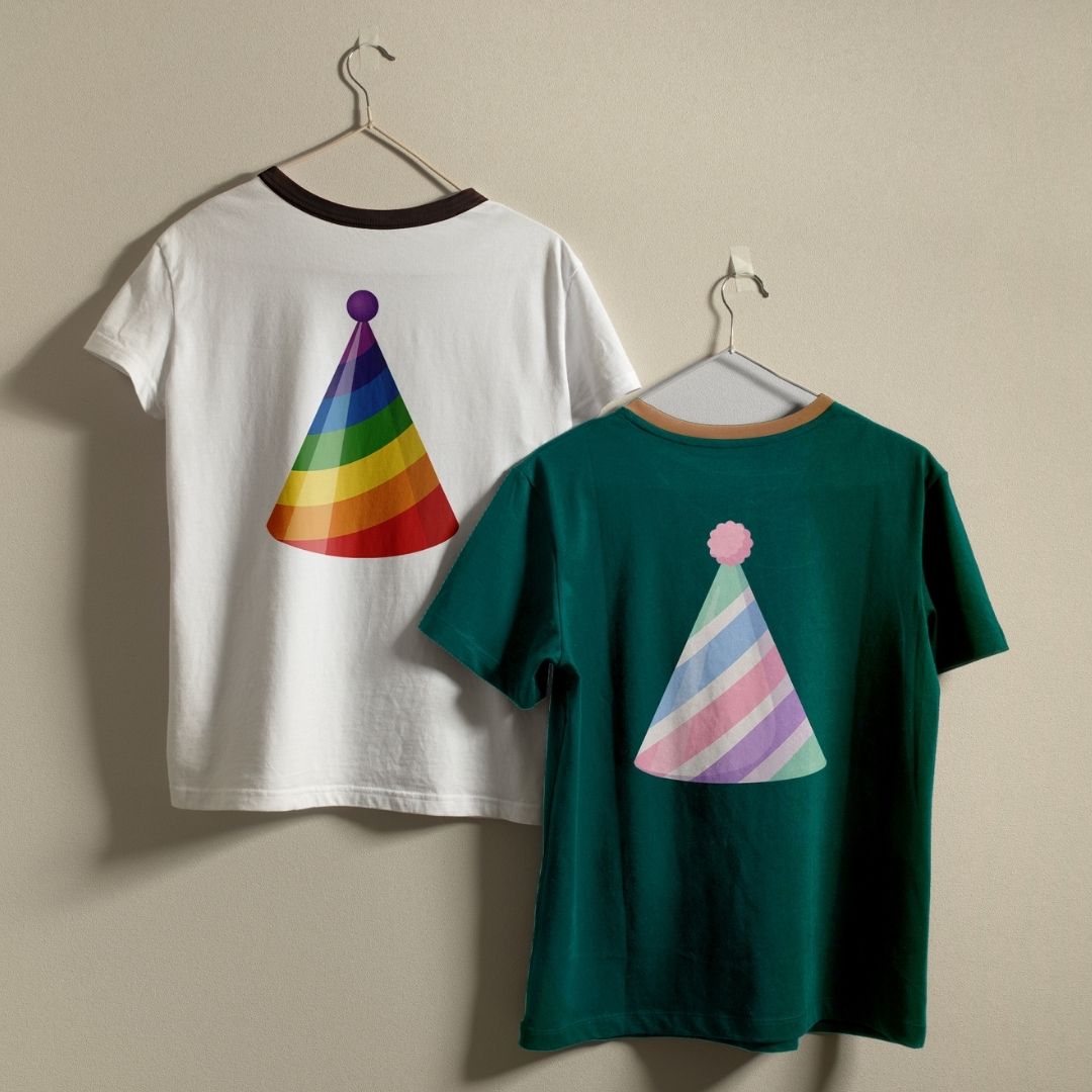 Two ringer t-shirts hanging on metal hangers against a plain beige wall. The left shirt is white with a black collar trim and features a colorful rainbow party hat printed on the back.