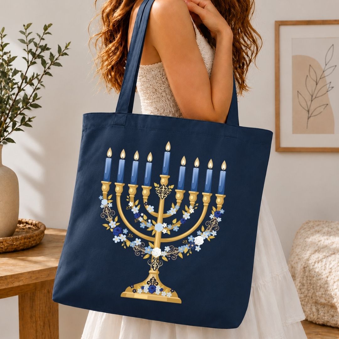 A woman holds a navy blue canvas tote bag featuring a large gold Hanukkah menorah design with blue candles and white floral accents, set against a warm, cozy indoor background.
