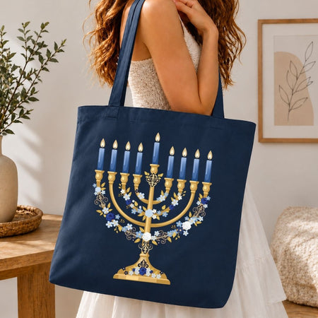 A woman holds a navy blue canvas tote bag featuring a large gold Hanukkah menorah design with blue candles and white floral accents, set against a warm, cozy indoor background.