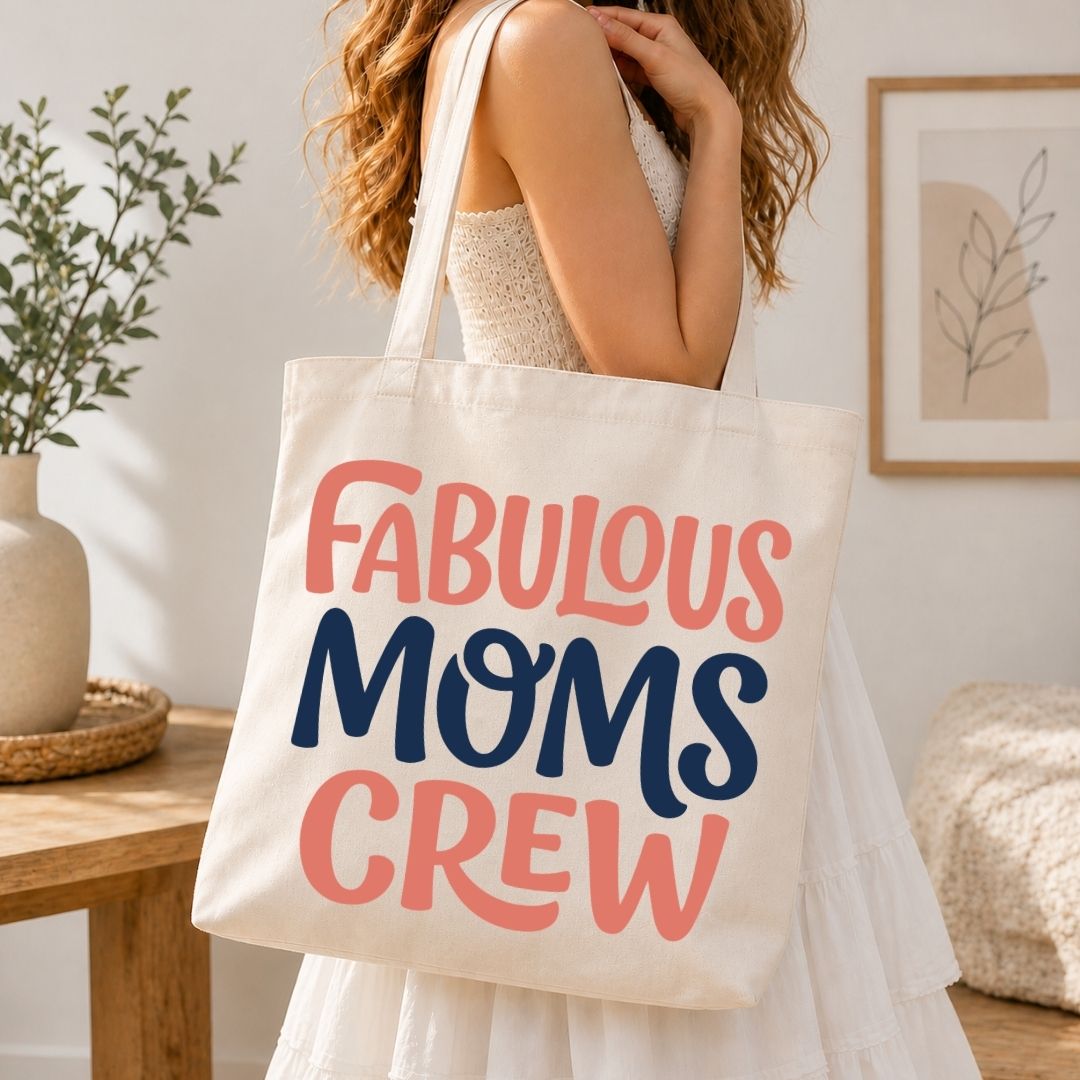 A woman in a white dress carries a cream canvas tote bag with the text 'FABULOUS MOMS CREW' in coral pink and navy blue lettering, set in a bright, modern room.