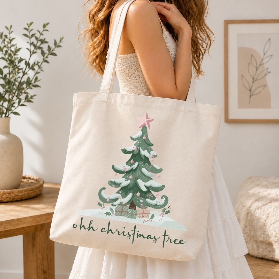 Woman holding a canvas tote bag printed with an "ohh christmas tree" typography, snow-covered tree, and present box holiday design.