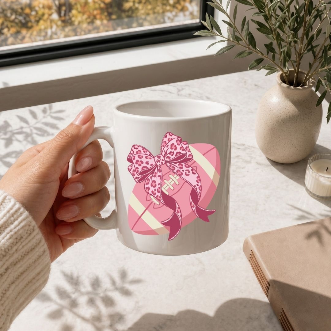 White mug with pink bow design held by a hand on a light surface with a window and plant in the background.