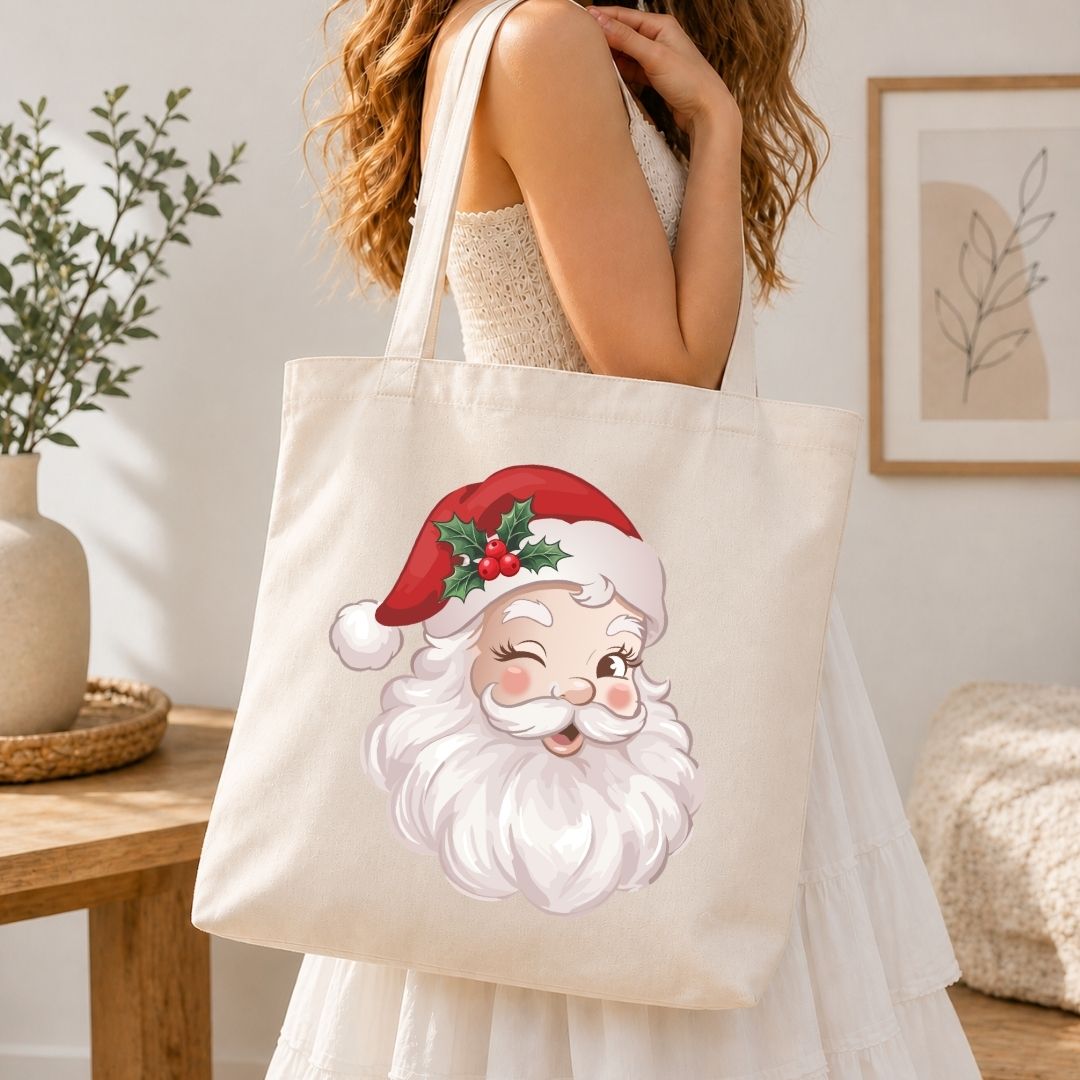 A woman in a white dress carries a cream canvas tote bag featuring a cute, winking cartoon Santa Claus face with a red hat and green holly leaves, set in a warm, modern room.