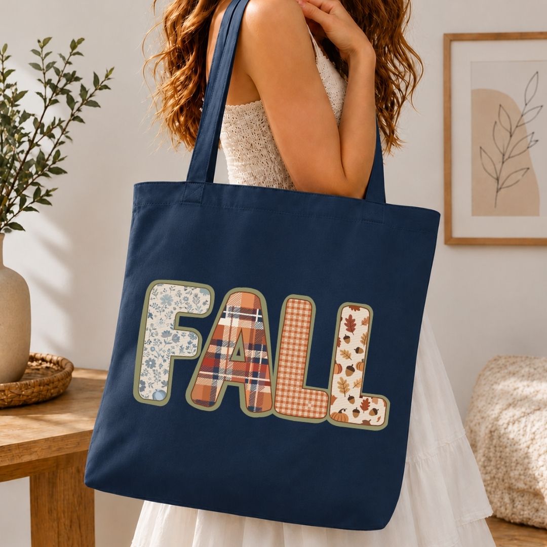 A navy blue canvas tote bag with the word 'FALL' spelled out in decorative autumn-themed patchwork letters, carried by a woman in a white dress in a warm, cozy indoor setting.