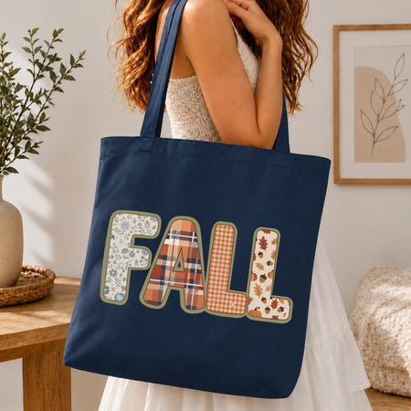 A navy blue canvas tote bag with the word 'FALL' spelled out in decorative autumn-themed patchwork letters, carried by a woman in a white dress in a warm, cozy indoor setting.