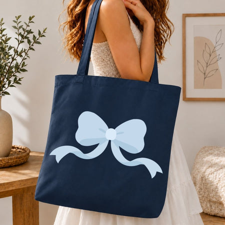 A navy blue canvas tote bag with a large light blue bow graphic, carried by a woman in a white dress in a warm, modern room setting.