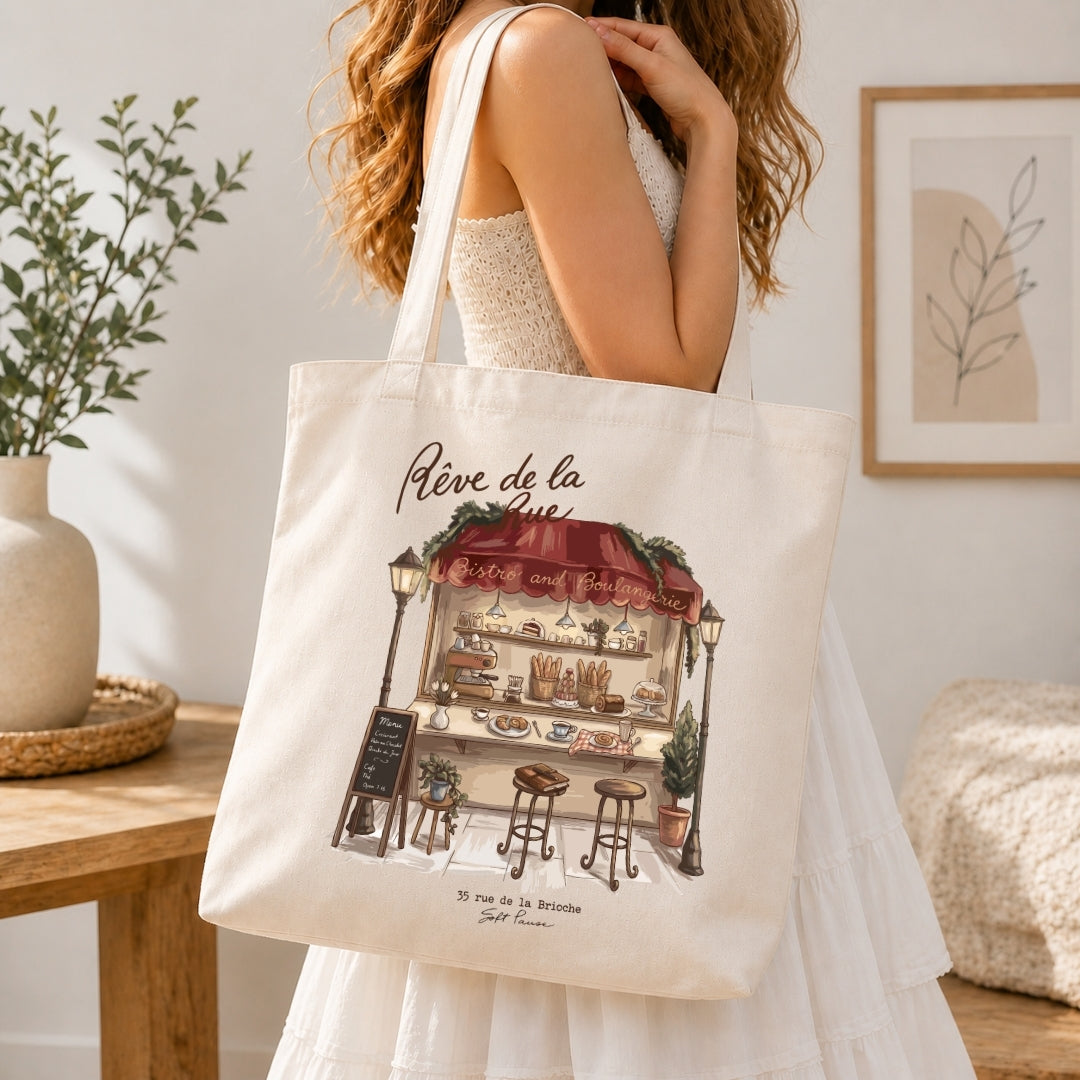 Person in a white dress carrying a canvas tote bag printed with the detailed "Rêve de la Rue" bakery stall graphic.