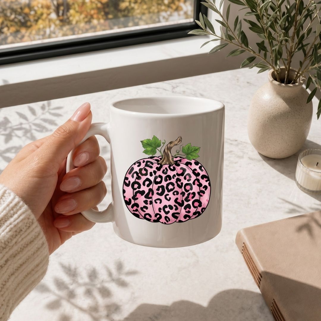 White mug with pink leopard print pumpkin design held by a hand on a light surface with a window and plant in the background.