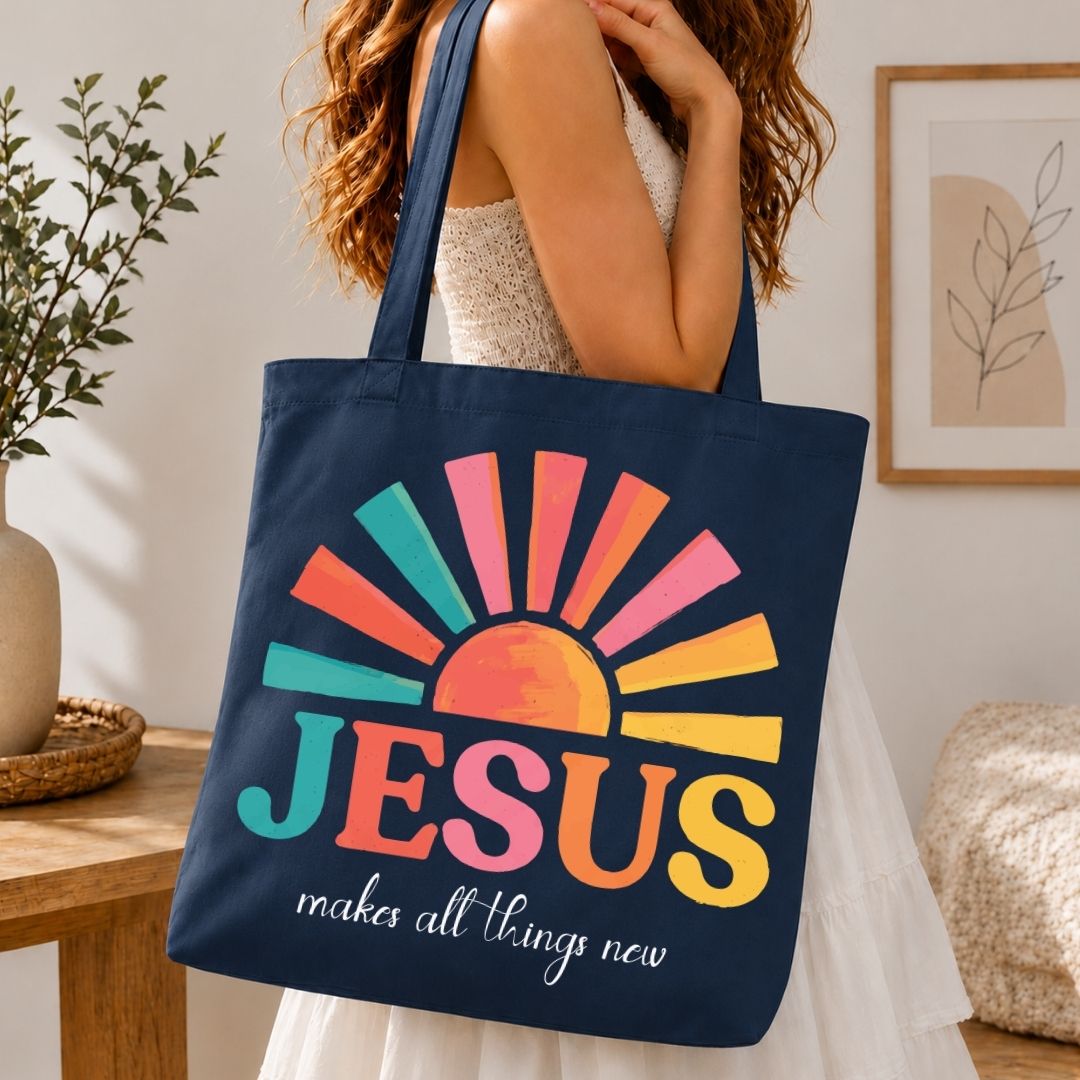 A navy blue canvas tote bag with a colorful sunburst graphic and the text 'JESUS makes all things new' in white script, carried by a woman in a white dress in a bright indoor setting.
