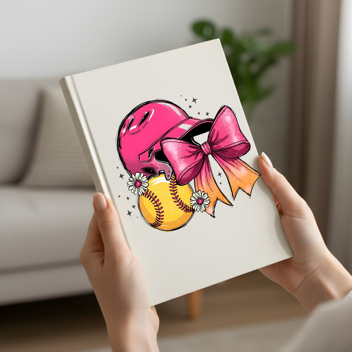 A person holds a cream-colored notebook or journal featuring a colorful graphic of a pink baseball helmet, a yellow softball, and a large pink bow on the cover, set against a blurred living room background.