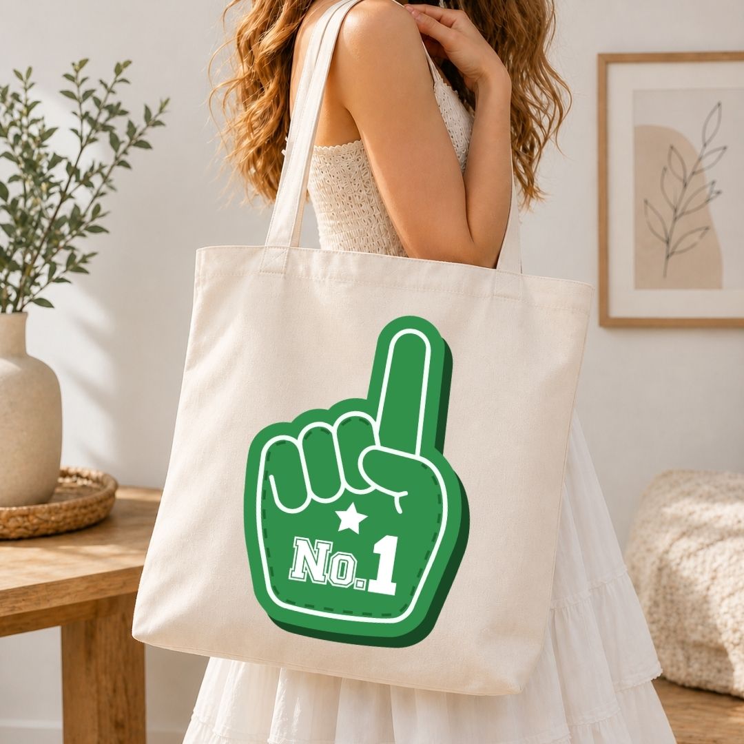 A woman wearing a white dress carries a cream canvas tote bag with a green foam finger graphic that says 'No. 1' in white text, set against a warm, softly lit indoor background.
