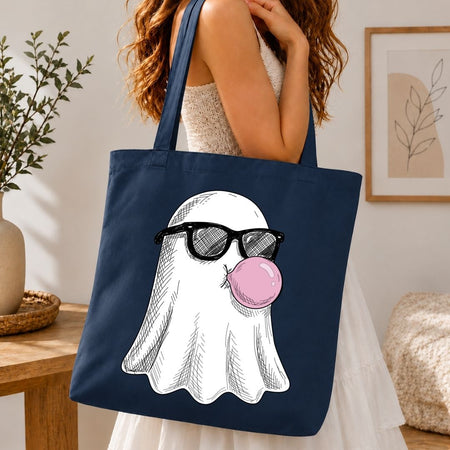 A navy blue canvas tote bag with a cartoon ghost wearing black sunglasses and blowing a pink bubblegum bubble, carried by a woman in a white dress in a cozy indoor setting.