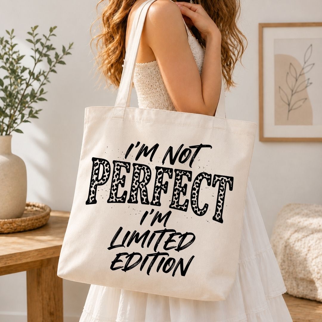 A woman in a white dress carries a large cream canvas tote bag with the text 'I'M NOT PERFECT I'M LIMITED EDITION' printed in black. The photo is taken in a bright, modern room with a wooden table, a potted plant, and framed wall art.