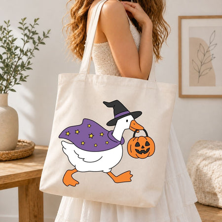 A woman wearing a white dress carries a cream canvas tote bag with a cartoon duck wearing a purple witch hat and cape while holding an orange jack-o'-lantern, in a bright, modern room.