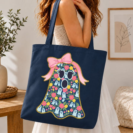 A navy blue canvas tote bag with a colorful Halloween ghost graphic, carried by a woman in a white dress in a cozy indoor setting.