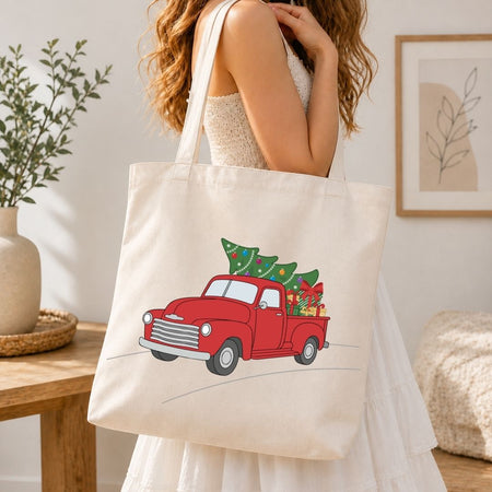 A woman wearing a white dress carries a cream canvas tote bag with a red vintage truck carrying a green Christmas tree printed on the front, set in a bright, modern indoor room.