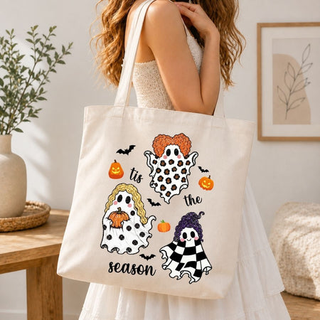 A woman wearing a white dress carries a cream canvas tote bag with a Halloween ghost design. The bag features two cute cartoon ghosts, pumpkins, bats, and the text 'it's the season'.