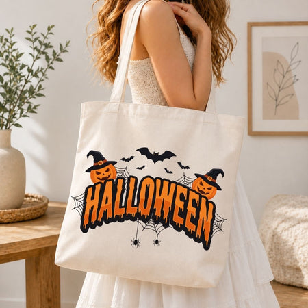 Woman holding a canvas tote bag printed with a "Halloween" typography, witch hats, pumpkins, and spiderweb holiday design.