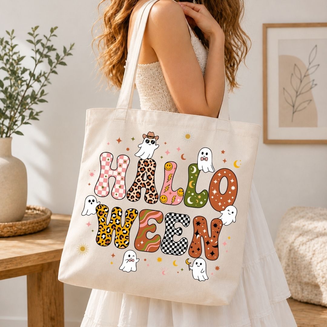 A woman wearing a white dress carries a cream canvas tote bag printed with the word 'HALLOWEEN' in patterned letters and small ghost illustrations, photographed in a softly lit indoor room.