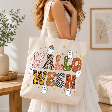 A woman wearing a white dress carries a cream canvas tote bag printed with the word 'HALLOWEEN' in patterned letters and small ghost illustrations, photographed in a softly lit indoor room.