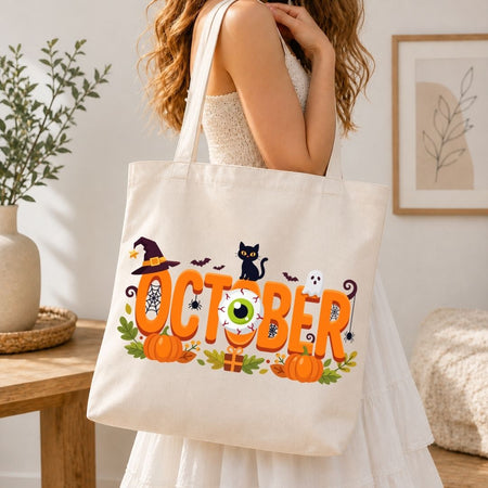 Woman holding a canvas tote bag printed with a spooky "October" Halloween graphic featuring an eyeball and pumpkins.