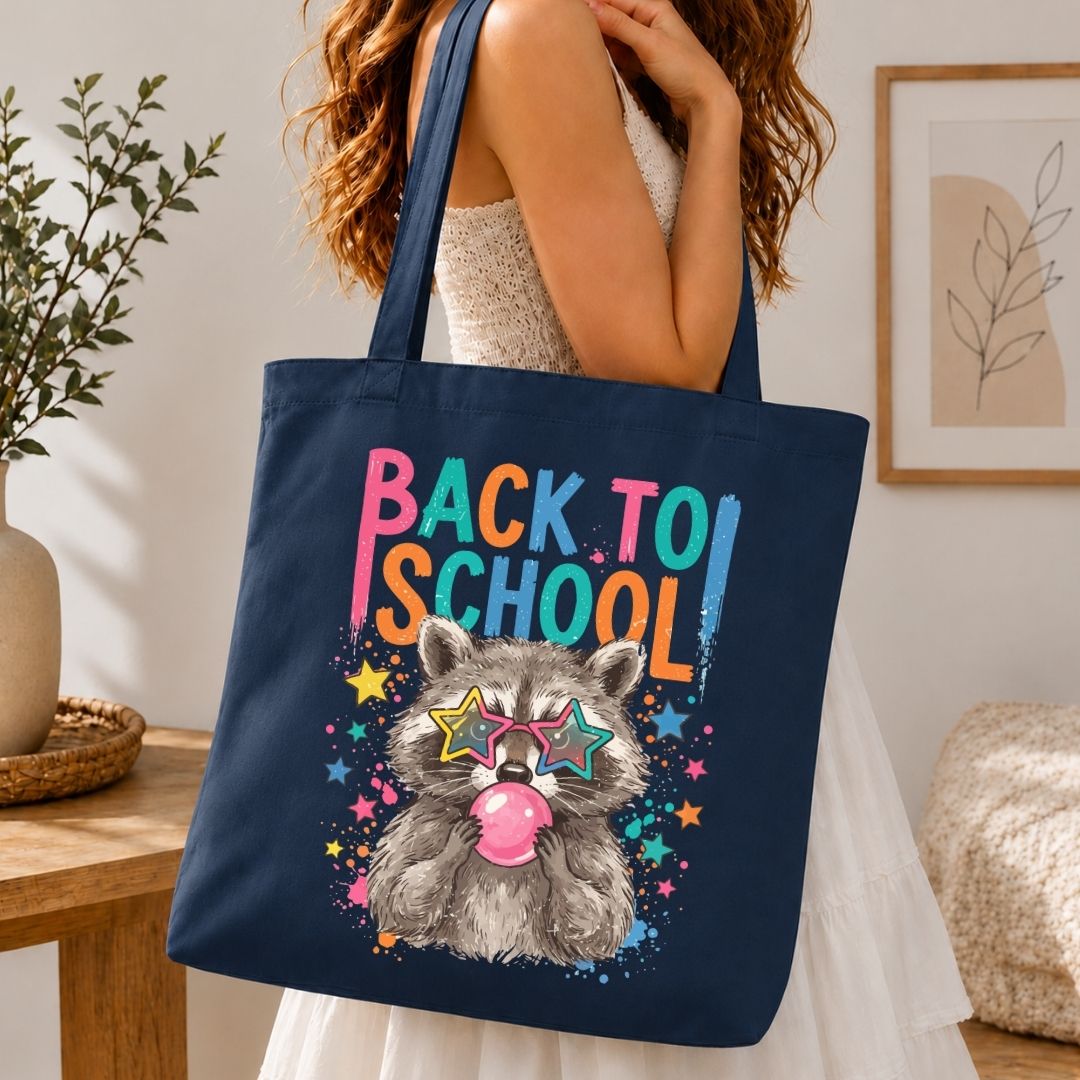 A navy blue canvas tote bag with a colorful 'Back to School' raccoon graphic, carried by a woman in a white dress in a cozy indoor setting.