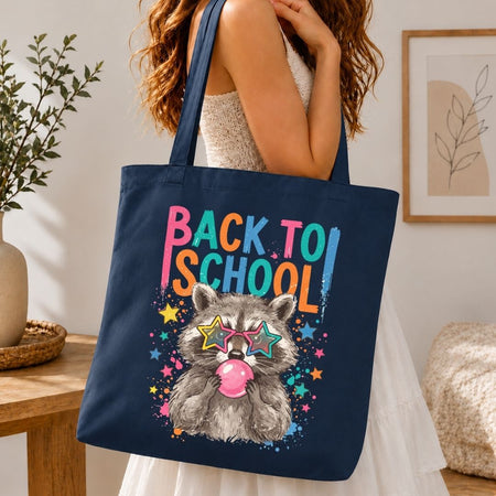 A navy blue canvas tote bag with a colorful 'Back to School' raccoon graphic, carried by a woman in a white dress in a cozy indoor setting.