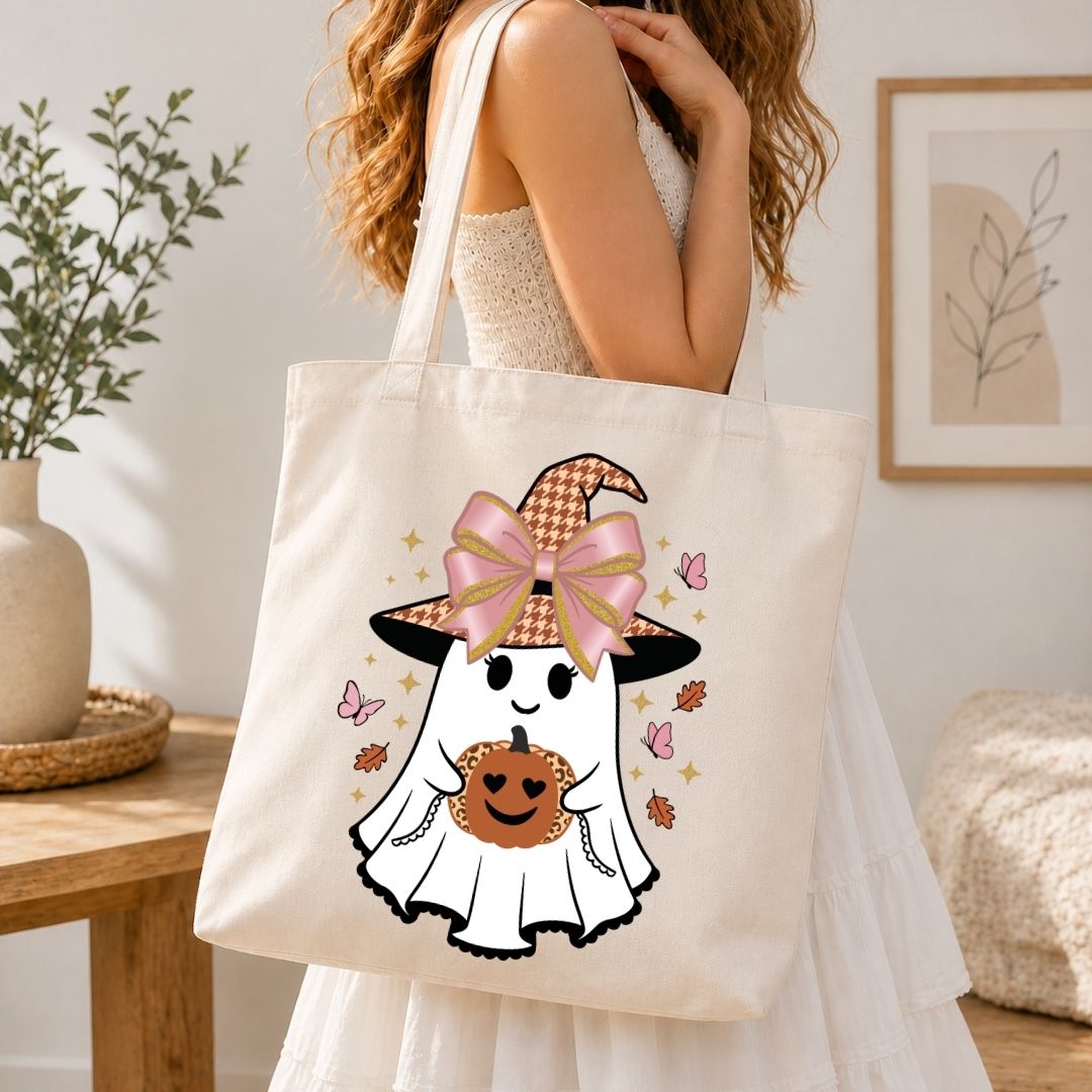 A woman wearing a white dress carries a cream canvas tote bag featuring a cute cartoon ghost in a pink bow witch hat holding a pumpkin. The scene is set in a warm, softly lit room with a wooden table, potted plant, and framed wall art in the background.